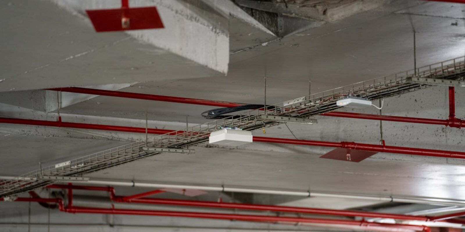 Red sprinkler pipes and cable trays running beneath a concrete parking structure ceiling